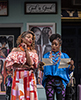 Madam Page wearing a bare-shoulder orange patterned blouse and orange and red  and white geometric patterned dress looks over at the two letters Madam ford, in baggy blue blouse, a dozen or more colorful bead necklaces hanging about her neck, and tight purple and black patterend yoga pants. Behind are signs of braided hair on the wall. and at the top a sign that says "God is Good all the time"